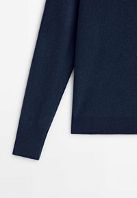 Manche et ourlet inférieur d’un pull en maille bleu marine à manches longues avec poignets côtelés sur fond blanc.