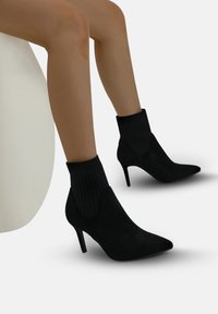 Stivaletti ankle in suede nera con una tomaia in tessuto a coste nera simile a una calza, punta affusolata e tacco alto sottile.