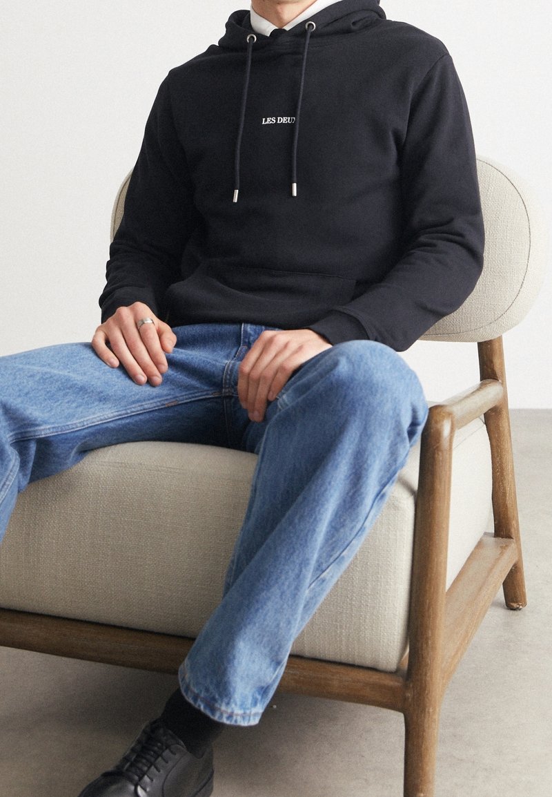 Czarna bluza z kapturem z logo "LES DEUX", z troczkami, noszona na koszuli z kołnierzykiem. Stylizowana z jasnoniebieskimi dżinsami i czarnymi butami na neutralnym krześle.