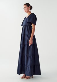 Námořnické maxi šaty s vrstveným designem, krátkými balónovými rukávy a čtvercovým výstřihem. Tkanina má hladkou texturu, doplněná o hnědé sandále.