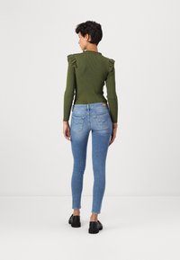 Γυναίκα που στέκεται με την πλάτη στραμμένη, φορώντας ένα ελαιόχρωμο τοπ με ρέουσες ώμους, ανοιχτό μπλε στενά τζιν και μαύρες loafers σε λευκό φόντο.