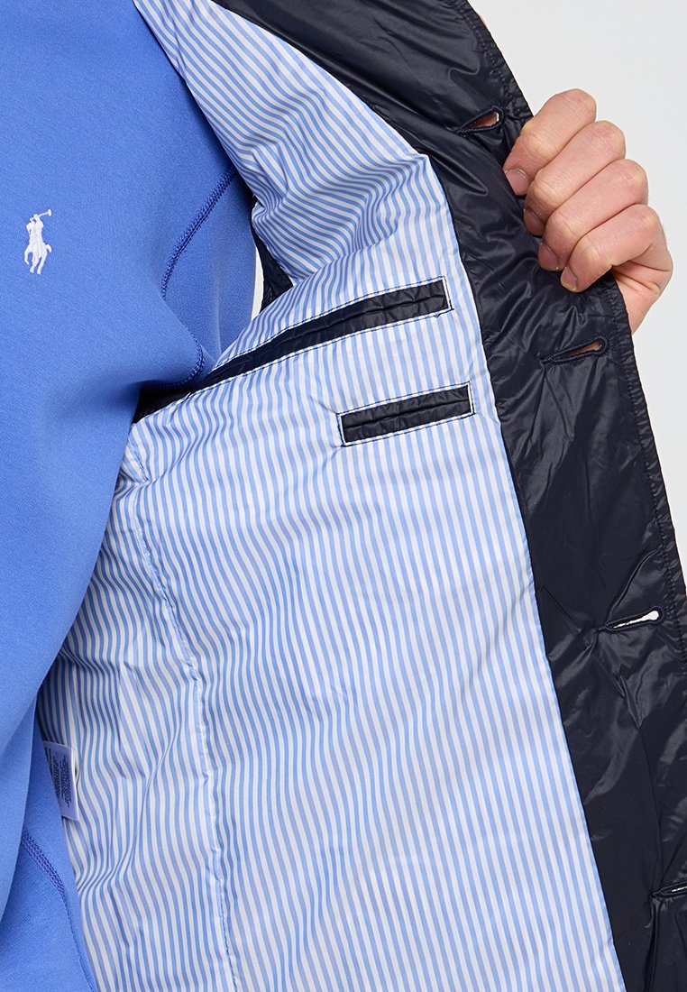 Granatowa kurtka z niebiesko-białym paskowanym wnętrzem, z zamykanymi na zamek kieszeniami i logo. Gładka zewnętrzna faktura z pikowanym wzorem.