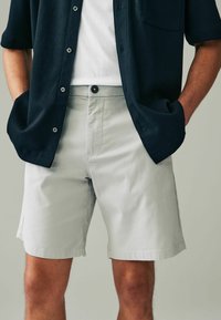 Helle graue Shorts aus Baumwollmischgewebe mit klassischem Schnitt, Knopfverschluss und Seitentaschen, kombiniert mit einem navyblauen Hemd mit Knöpfen.