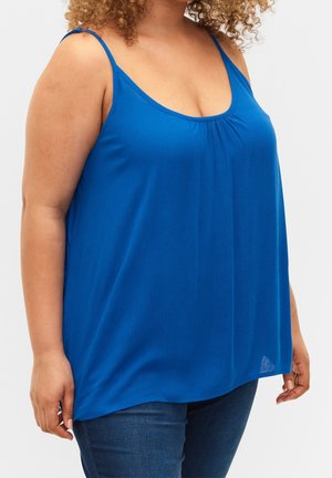 Femme aux cheveux bouclés portant un haut bleu ample sans manches et un jean bleu foncé, debout devant un fond uni.