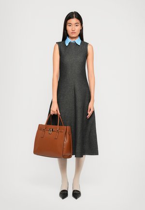 Woman in sleeveless gray dress with blue collar holds large brown leather handbag, wearing white stockings and black pointed shoes.