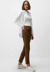 Pantalons à imprimé léopard marron avec une coupe slim, associés à un haut blanc ajusté et une chemise blanche à boutons surdimensionnée. Des baskets blanches complètent le look.