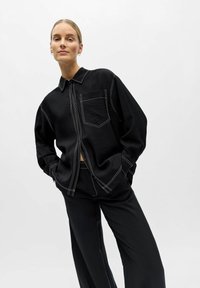 Femme portant une chemise noire ample et un pantalon avec des coutures contrastantes blanches, debout les mains dans les poches devant un fond blanc.