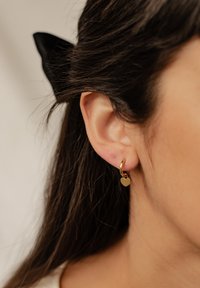 Orecchino a cerchio in oro con un piccolo ciondolo a forma di cuore, caratterizzato da una finitura liscia e una superficie riflettente, indossato su un orecchio su uno sfondo neutro.