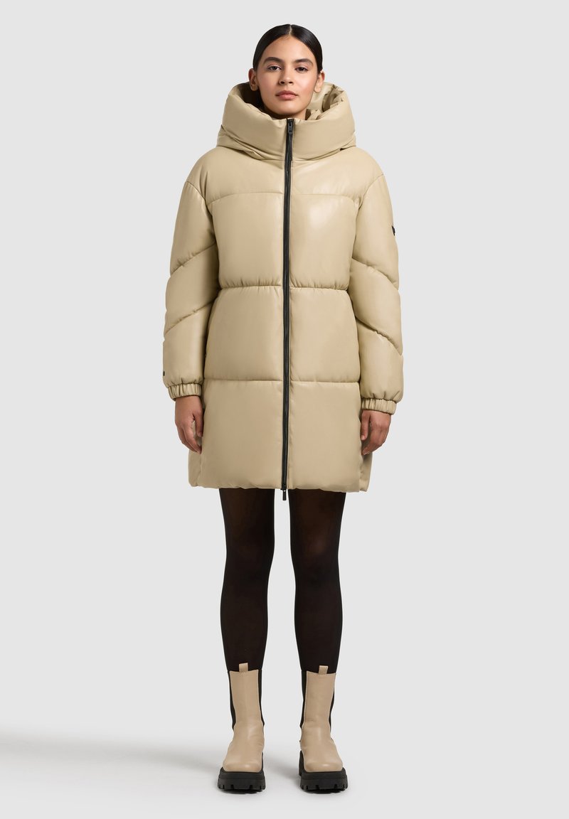 Abrigo acolchado beige con cuello alto y capucha, que cuenta con una cremallera central, mangas elásticas y una textura brillante. Combinado con botas de tobillo.