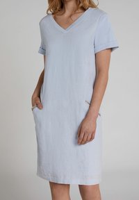 Lichtblauwe linnen jurk met een V-hals, korte opgerolde mouwen en twee zakken met subtiele metallic accenten. Rechte snit.