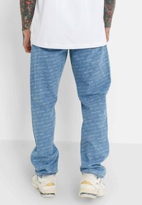Hombre con jeans de un azul claro con un patrón repetido de texto "SIXTH JUNE", zapatillas blancas y una camiseta blanca, visto desde atrás por debajo de la cintura.