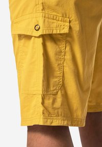 Žluté cargo šortky vyrobené z lehkého materiálu, s boční kapsou a černým knoflíkem a viditelným šitím podél lemu.