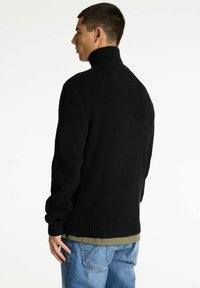 Schwarzer gerippter Rollkragenpullover mit eng anliegenden Ärmeln, hohem Kragen und strukturiertem Design, getragen über einer grünen Schicht und kombiniert mit blauen Jeans.