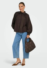 Camicia marrone con bottoni e maniche a sbuffo, abbinata a jeans blu a gamba dritta e una borsa a tracolla nera trapuntata. Tacchi slingback marroni completano il look.