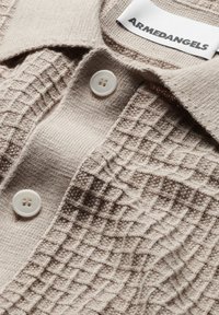 Polo shirt beige a maglia testurizzata con colletto a costine, tre bottoni bianchi e un motivo in rilievo sul davanti. L'etichetta riporta "ARMEDANGELS."