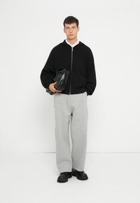 Felpa nera con zip a coste, abbinata a pantaloni sportivi grigi e sneakers nere. Il modello tiene una borsa di pelle nera, in piedi su uno sfondo bianco.