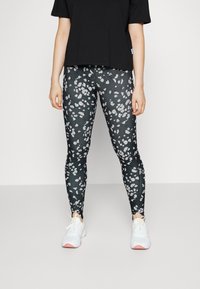 Černé legíny s šedým leopardím vzorem, kombinované s volným černým tričkem a bílými sportovními botami se žlutými akcenty.