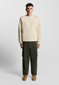 Felpa color crema con collo a giro, abbinata a pantaloni cargo verde oliva e scarpe slip-on color beige, indossata da una persona in piedi.