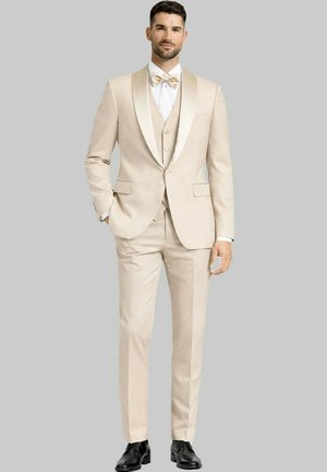 Homme en costume trois pièces beige avec nœud papillon et chaussures habillées noires, debout devant un fond uni gris clair.