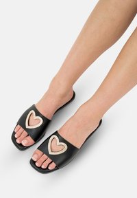 Sorte flip-flops med et stort hjerteudskæring, indvendigt i beige og med rhinsten-accenter. Kvadratisk tå og flad sål design.