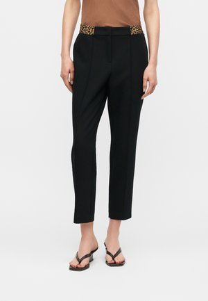 Woman wearing black tailored ankle-length pants with leopard print waistband panels, brown sleeveless top, and black heeled sandals.