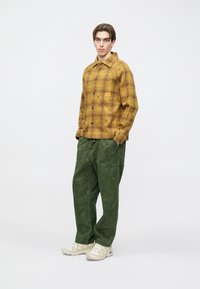 Żółta flanelowa koszula w kratę, z kieszenią na piersi. Luźne zielone spodnie z fakturowanym wzorem i sznurkiem w pasie. Jasne sneakersy.