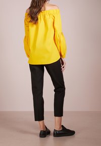 Žena v žluté blůze s odhalenými rameny, černých zkrácených kalhotách a černých teniskách stojící proti jednobarevnému pozadí, otočená zády.