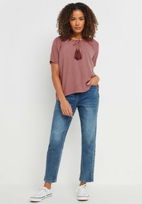 Różowa bluzka z krótkim rękawem i okrągłym dekoltem, ozdobiona frędzlami, zestawiona z jasnoniebieskimi spodniami straight leg i białymi trampkami.