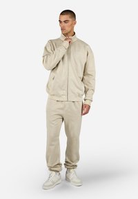 Beige krūšturis ar rāvējslēdzēju un augstu apkakli, apvienots ar atbilstošām sporta biksēm ar auklu. Abiem ir mīksta, strukturēta auduma. Balti sporta apavi pabeidz izskatu.