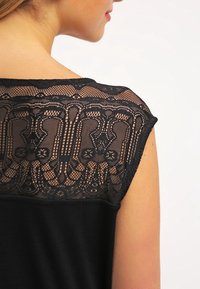 Černý top s krajkovým zadním dílem s jemnými vzory, krátkými rukávy a hladkým tělem z látky. Elegantní krajková výzdoba je výrazná.