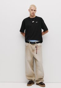 Sort bomulds T-shirt med logo, lys beige baggy jeans, sort bælte med hardware og mørkebrune sko. Modellen står mod en hvid baggrund.