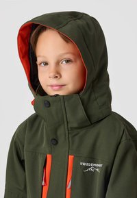 Olivgrön jacka med fodrad huva, med ljusa orange detaljer, stora knappar och en logotyp. Texturerat tyg med synliga sömmar.