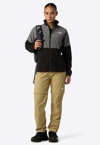 Černá a šedá fleecová bunda na zip s kostkovaným vzorem, v kombinaci s khaki kalhotami na zip a černými turistickými botami, držící v ruce láhev s vodou.