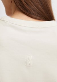 Helle beige Baumwoll-T-Shirt mit rundem Ausschnitt und dezentem gesticktem Logo-Detail auf dem oberen Rücken. Glatte Textur.