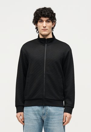 Jonge man met krullend donker haar, gekleed in een zwart jack met ritssluiting met een gestructureerd patroon en lichtblauwe spijkerbroek, staand tegen een witte achtergrond.