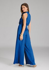 Gioco intero blu royal con gambe larghe, tessuto texturizzato, design senza maniche e dettaglio legato sul retro, abbinato a accessori dorati.