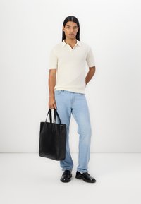 Kortermet kremfarget polo skjorte, lyseblå jeans, svart lær totebag med to håndtak, og svarte sko, satt mot en ensfarget bakgrunn.