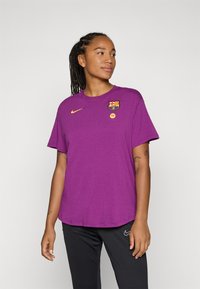 Ljubičasta Nike majica s okruglim izrezom, kratkim rukavima i logotipom Barcelone. Mekana tekstura s opuštenim krojem. Crne hlače ispod.
