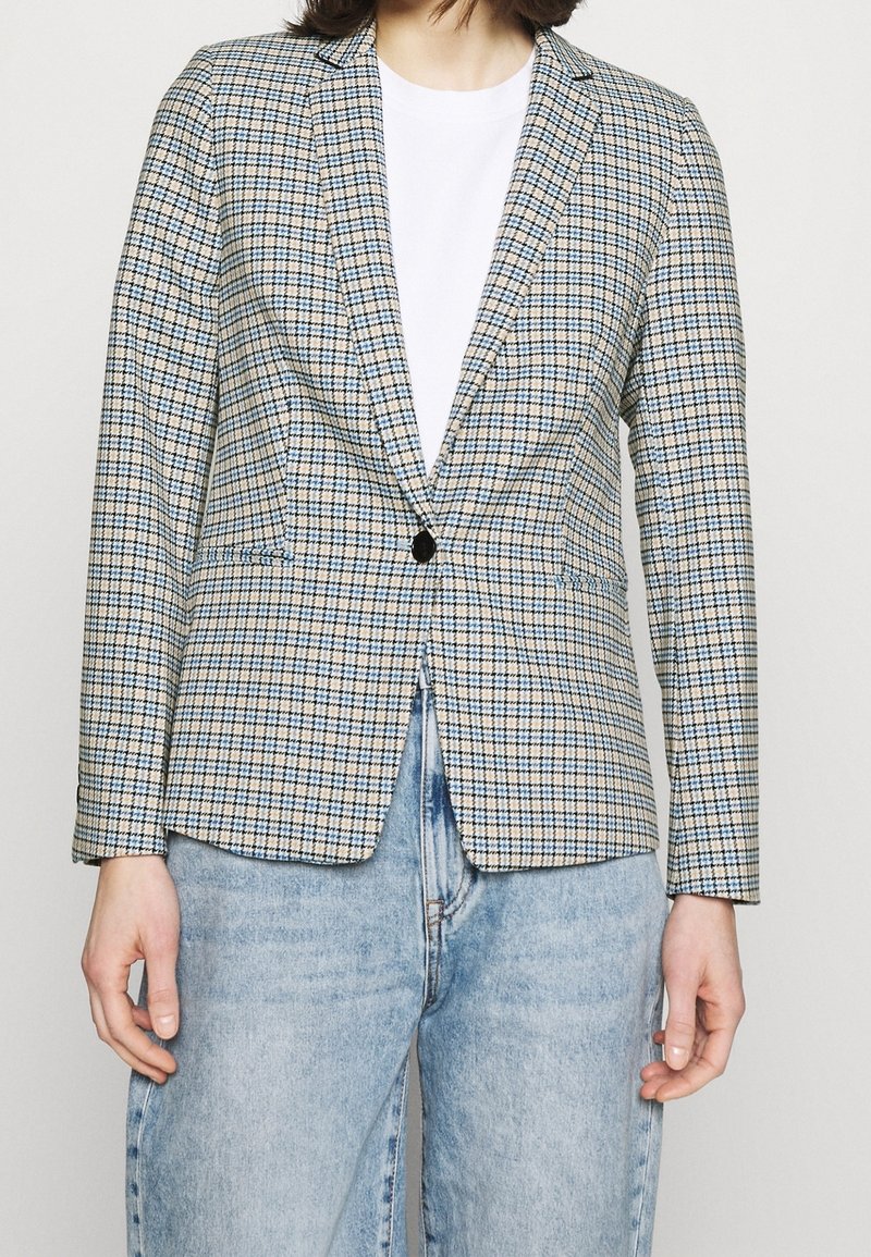Blazer a quadri in beige chiaro, blu e nero con rever affilati, chiusura con un solo bottone e due tasche laterali, abbinato a una maglietta bianca.