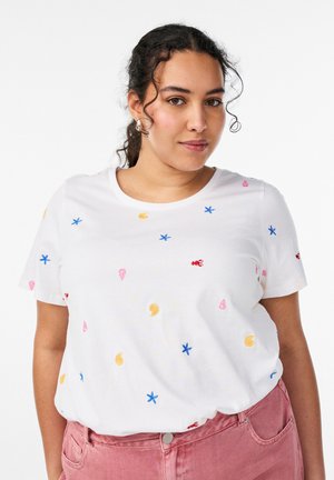 Ung kvinde med krøllet hår iført sølv hoops-øreringe, en hvid T-shirt med farverigt havtema broderi og lyserøde bukser.