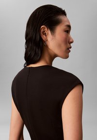 Svart figursydd klänning med korta ärmar, slät textur och subtil detalj med dragkedja baktill. Modellen har blött hår och bär kreolörhängen.