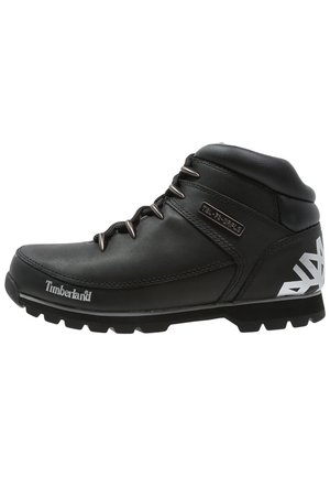 Czarny trekkingowy skórzany but Timberland z metalowymi oczkami, białym logo z boku i na pięcie oraz grubą, teksturowaną podeszwą.