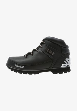 Czarny trekkingowy skórzany but Timberland z metalowymi oczkami, białym logo z boku i na pięcie oraz grubą, teksturowaną podeszwą.