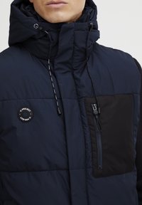 Mornarica prsluk s kumulacijom crne boje na džepu, zatvaranje na zip, i podesiva kapuljača. Sadrži okrugli logo i naglašene detalje na vezici. Glatka, quilted tekstura.