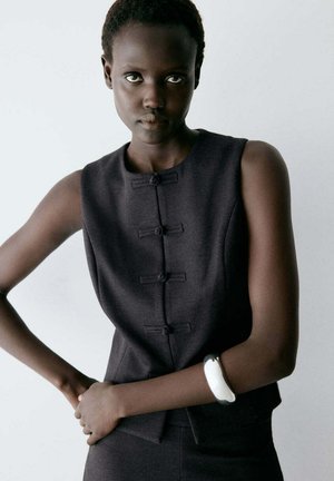 Femme aux cheveux courts portant un haut noir sans manches avec des boutons, posant les mains jointes, portant un bracelet blanc à son poignet gauche.