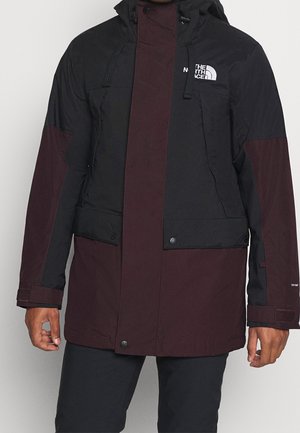 Mężczyzna w czarnej i ciemnoczerwonej kurtce z kapturem North Face z suwakiem, zapinanymi kieszeniami na guziki oraz regulowanymi paskami przy mankietach.