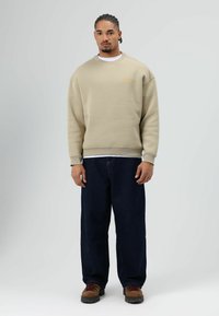 Beige sweatshirt met geribbelde manchetten en zoom, gecombineerd met losse, donkerblauwe jeans. Bruine laarzen met zwarte zolen, neutrale achtergrond.
