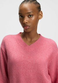 Rosa V-Ausschnitt-Pullover aus weichem Strickmaterial mit lockerem Schnitt. Verfügt über leicht abfallende Schultern und gerippte Textur am Ausschnitt.