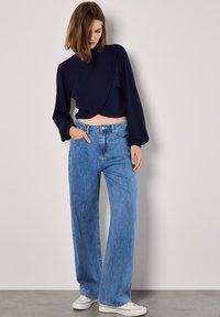 Marineblaues, geripptes, langärmliges Cropped Top mit hochgeschlossenem Kragen, kombiniert mit hellblauen, weit geschnittenen Jeans. Das Model trägt weiße Sneakers.