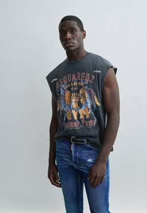 Νεαρός άνδρας στέκεται με σοβαρή έκφραση, φορώντας αμάνικο γυμναστηριακό μπλουζάκι με σχέδιο και μπλε τζιν, μπροστά σε απλό ανοιχτό φόντο.